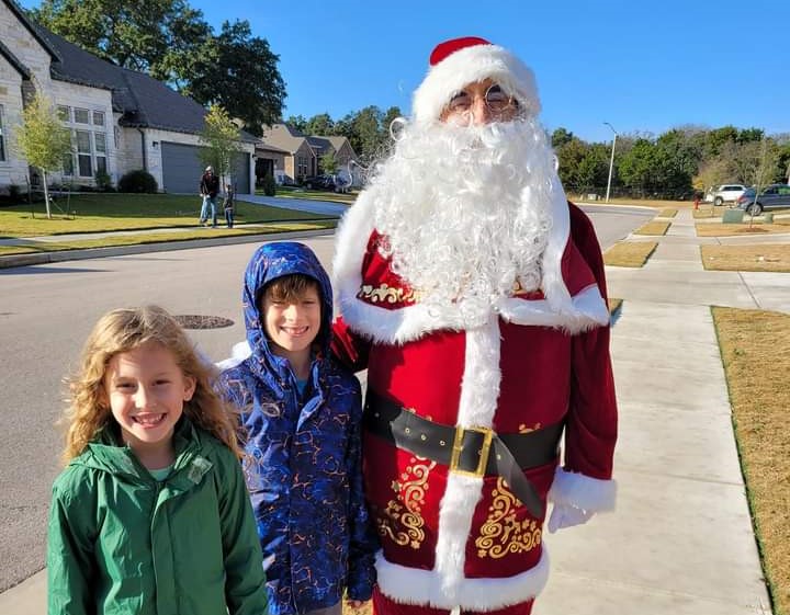 An image of a person in a red and white Santa suit with two children. The costumed person and the children are standing on Split oak Drive facing the cul-de-sac.